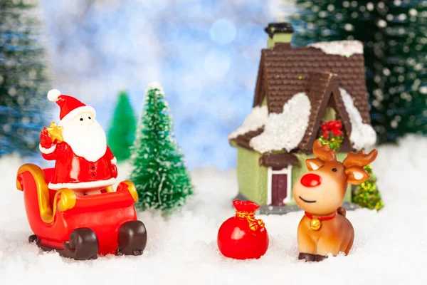 Ano Novo cartão postal cumprimentando. Um Pai Natal de brinquedo está num trenó de Natal. Nas proximidades há uma casa de brinquedos, um boneco de neve, um veado. A nevar. Em torno de abetos de brinquedo — Fotografia de Stock