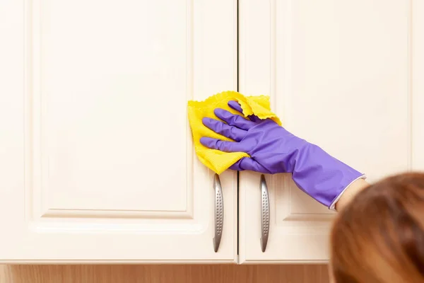 Mano femenina en un guante de goma púrpura limpia la puerta del gabinete de cocina beige. Trapo amarillo en la mano — Foto de Stock