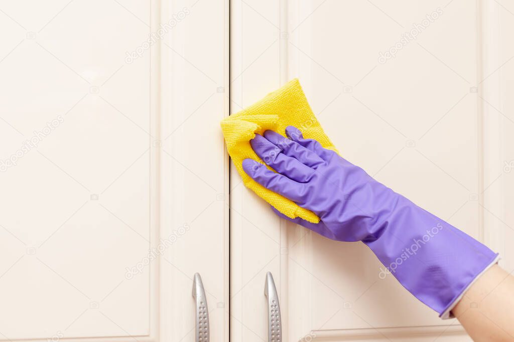 female hand in a purple rubber glove wipes the beige kitchen cabinet door. Yellow rag in hand
