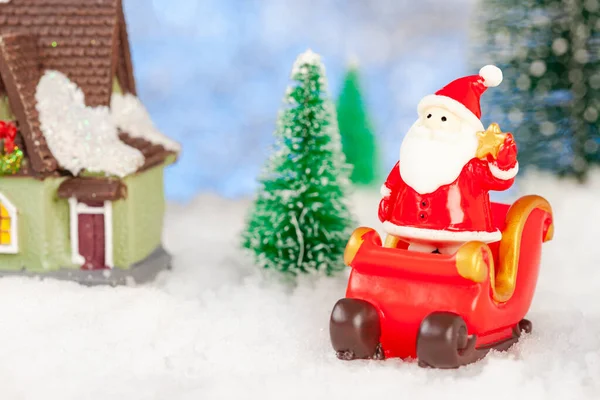Πρωτοχρονιάτικη κάρτα. Ένα παιχνίδι Σάντα Κλάους στέκεται σε ένα Χριστουγεννιάτικο έλκηθρο. Εδώ κοντά είναι ένα σπιτάκι με παιχνίδια, ένας χιονάνθρωπος, ένα ελάφι. Χιονίζει. Γύρω από έλατα παιχνιδιών — Φωτογραφία Αρχείου