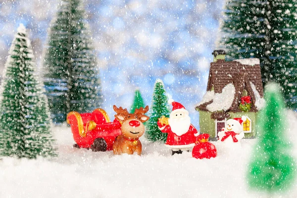 Ano Novo cartão postal cumprimentando. Um Pai Natal de brinquedo está num trenó de Natal. Nas proximidades há uma casa de brinquedos, um boneco de neve, um veado. Neve e brinquedo abetos ao redor — Fotografia de Stock