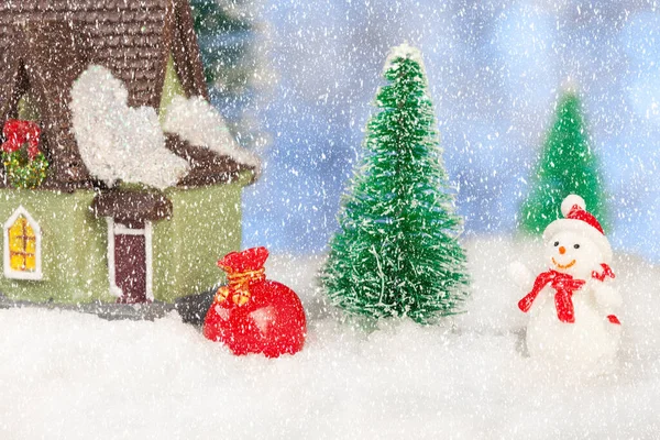 Boneco de neve de brinquedo com um saco de presentes perto de uma pequena casa, cartão de felicitações de Natal — Fotografia de Stock