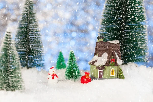 Boneco de neve de brinquedo com um saco de presentes perto de uma pequena casa, cartão de felicitações de Natal — Fotografia de Stock