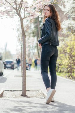 Siyah kotlu ve deri ceketli bir kız sakura ağacının yanındaki kaldırımda yürüyor.