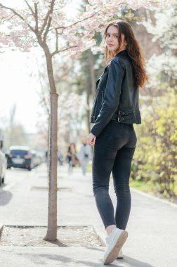 Siyah kotlu ve deri ceketli bir kız sakura ağacının yanındaki kaldırımda yürüyor.