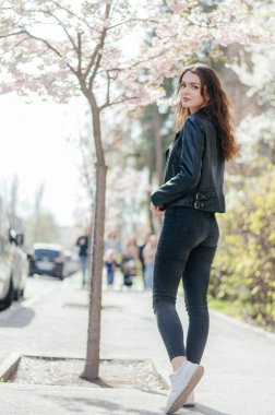 Siyah kotlu ve deri ceketli bir kız sakura ağacının yanındaki kaldırımda yürüyor.