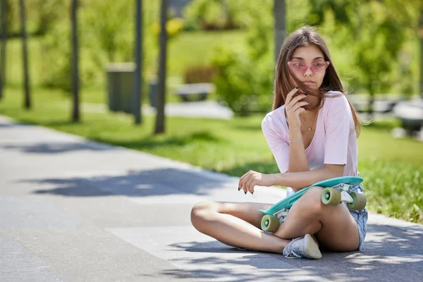 Красивая девушка-скейтбордистка со скейтбордом сидит на открытом воздухе — стоковое фото