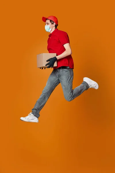 Delivery man in face mask holding cardboard box and jumping — Stock Photo, Image