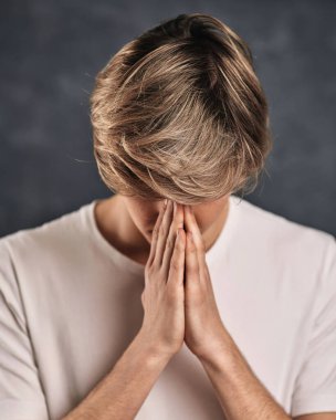 Adam dünya barışı için dua ediyor. Gri arka planda yoga jestinde el ele tutuşan şık saç kesimi olan genç adam.