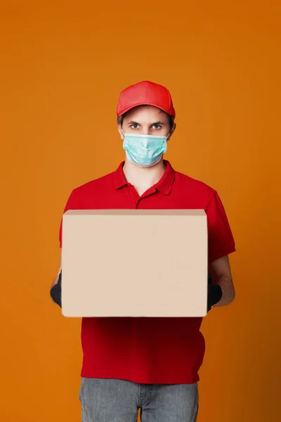 Delivery man in red cap, empty t-shirt, face mask, gloves, empty cardboard box isolated on orange background