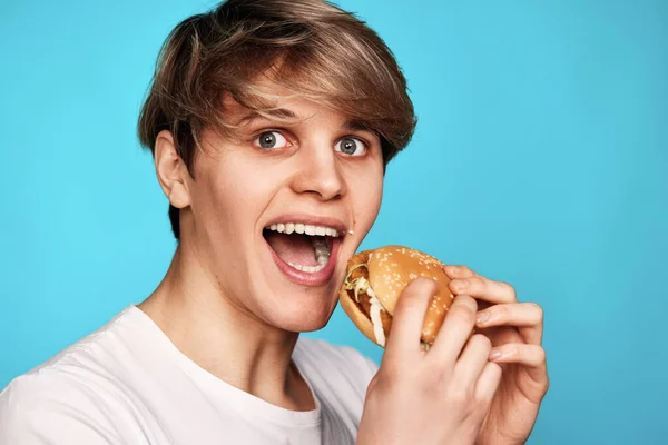 拿着美味汉堡包的年轻人被蓝色背景隔离了 那家伙吃快餐 — 图库照片