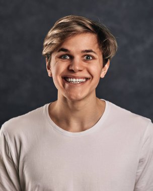 Beyaz tişörtlü, gri arka planda gülümseyen genç yakışıklı adam. İnsan duyguları, yüz ifadeleri.