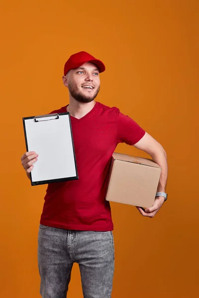 Delivery man in red cap, t-shirt holding clipboard and empty cardboard box isolated on orange background