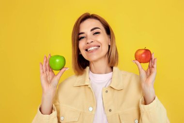 Sarı arka planda yeşil ve kırmızı elma tutan gülümseyen sarışın kadın. sağlıklı yiyecekler