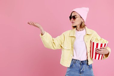 Güneş gözlüklü genç hippi kadın, elinde patlamış mısır kovası olan şapka pastel pembe arka planda fotokopi alanını işaret ediyor..