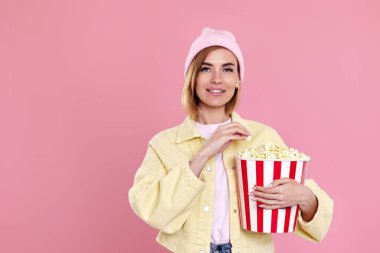 Şapkalı neşeli genç kız film izliyor, elinde pastel pembe arka planda izole edilmiş patlamış mısır kovası var..