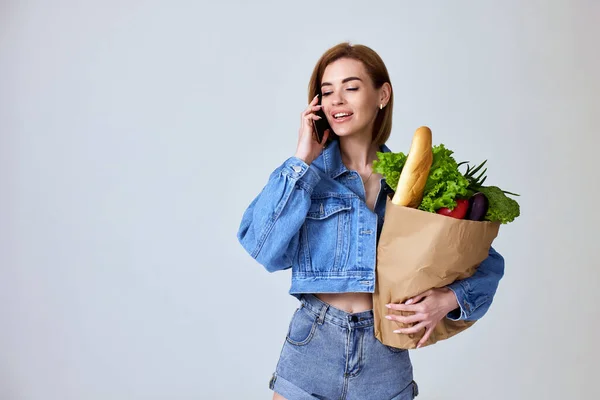 beautiful vegetarian woman holding shopping eco-friendly paper bag full of groceries and using phone on studio gray background. copy space.