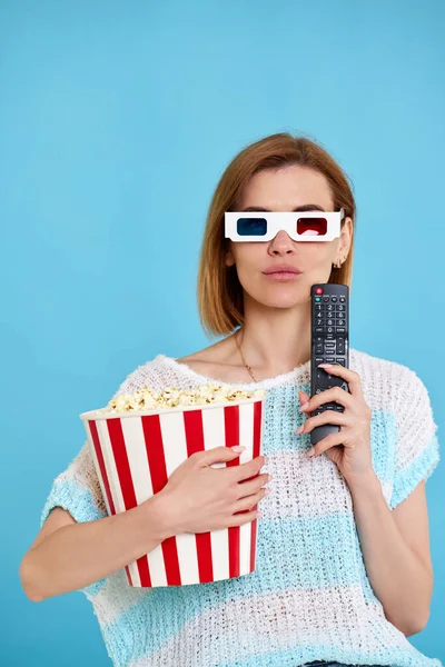 beautiful woman in 3d glasses using tv control, watching movie film and holding bucket of popcorn on blue background