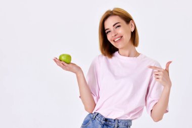 Gülümseyen güzel kız yeşil elma tutuyor ve beyaz arka planda izole edilmiş bir parmağı işaret ediyor.