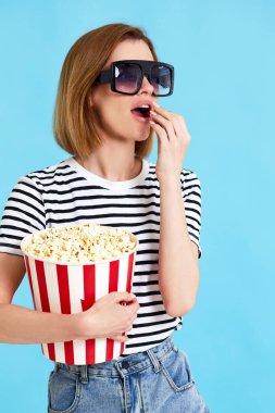 Güzel kız film izliyor ve mavi arka planda izole edilmiş patlamış mısır yiyor.