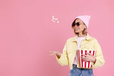 Güneş gözlüklü neşeli mutlu kadın, elinde pastel pembe arka planda izole edilmiş patlamış mısır kovası tutan şapka..