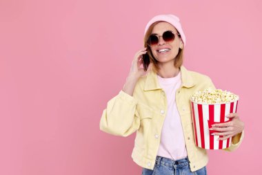 Güneş gözlüklü, şapkalı, patlamış mısırlı genç bir kadın telefonda pastel pembe arka planda konuşuyor. Boşluğu kopyala