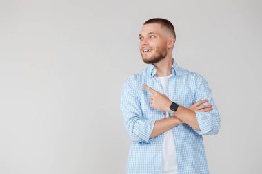 Genç yakışıklı sakallı adam, gri arka planda izole edilmiş bir ürünü sunmak için kenarı işaret ediyor.