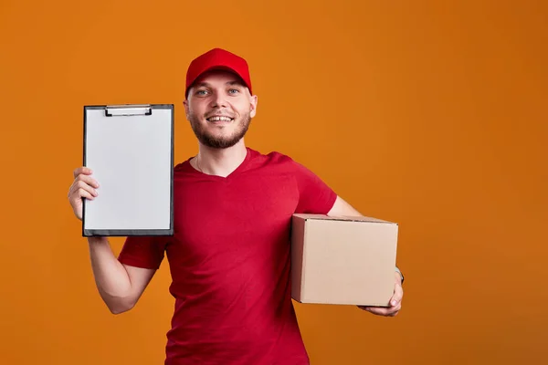 friendly delivery man in red cap, t-shirt holding clipboard and empty cardboard box isolated on orange background