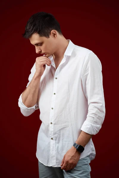 thoughtful man in white shirt holding arms folded, touching chin with hand and looking down on red background