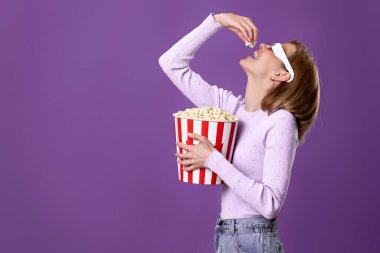 Üç boyutlu gözlüklü beyaz kadın film izliyor, mor arka planda izole edilmiş patlamış mısır yiyor.