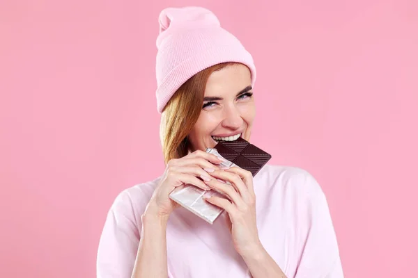 young happy woman in casual t-shirt biting sweet chocolate bar and looking at camera isolated on pastel pink background