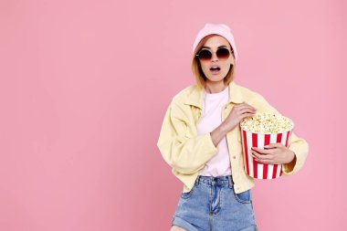 beautiful amazed woman in sunglasses with bucket of popcorn watching exciting movie on pink background. copy space