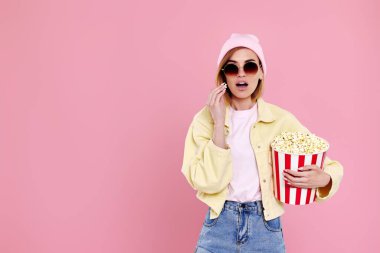 amazed hipster girl in sunglasses with bucket of popcorn watching exciting movie on pastel pink background. copy space