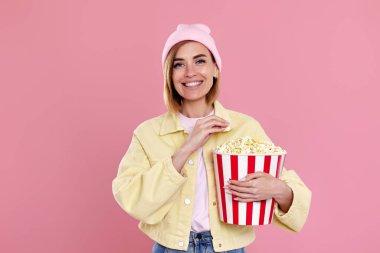 smiling girl in hat watching movie film, holding bucket of popcorn isolated on pastel pink background.