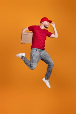 Delivery man in red cap, t-shirt holding cardboard box and jumping isolated on orange background