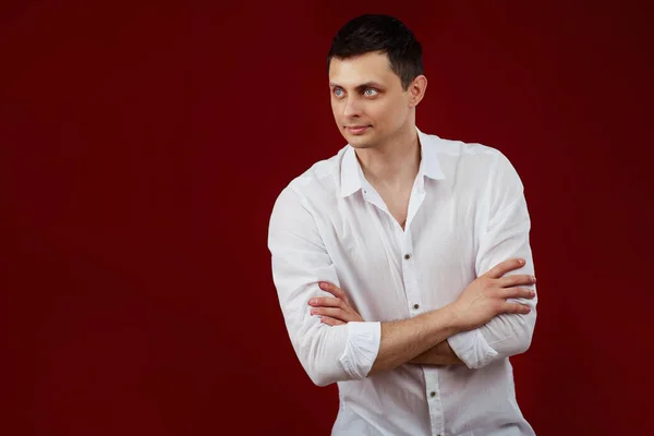 smiling young man in white shirt posing with crossed arms and looking at the side on red background