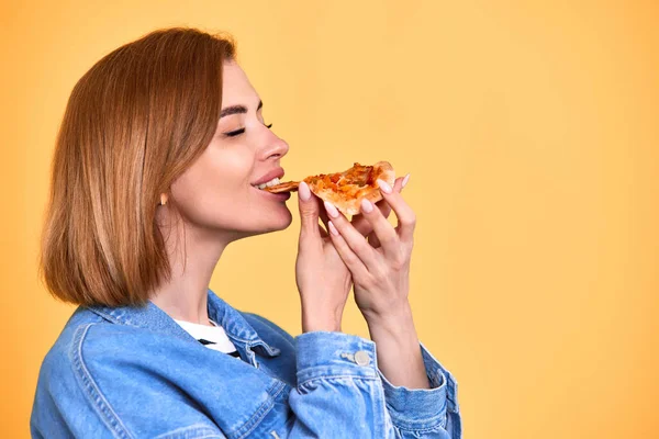 Молода Жінка Їсть Задоволенням Шматочок Піци Виглядає Захваті Жовтому Фоні — стокове фото