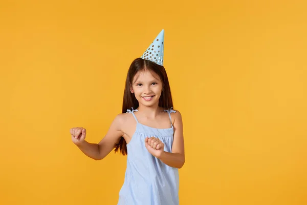 happy birthday child girl in blue dress and party hat dancing on yellow background