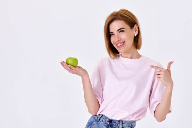 Portrait of a cheerful woman in pink t-shirt holding green apple and pointing finger isolated on white background. copy space