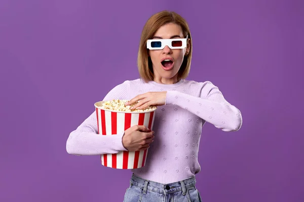scared terrified woman in 3d glasses watching movie film, holding bucket of popcorn on purple background