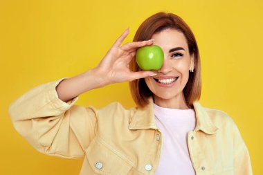 Portrait of smiling cheerful woman covers one eye with fresh green apple isolated on yellow background. copy space