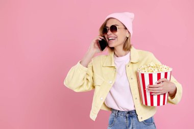 Güneş gözlüklü, şapkalı, patlamış mısırlı genç bir kadın telefonda pastel pembe arka planda konuşuyor. Boşluğu kopyala