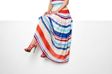 female legs wearing maxi striped summer dress and pink high heels shoes sitting on white bench.