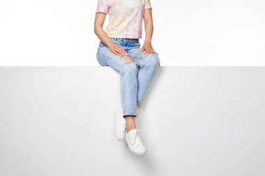 female legs in jeans and white sneakers sitting on white bench