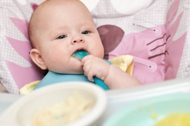 cute baby girl eating with spoon on baby chair in kitchen