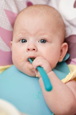 cute baby girl eating with spoon on baby chair in kitchen