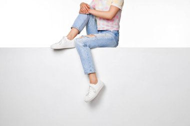 female legs in jeans and white sneakers sitting on white banner. copy space
