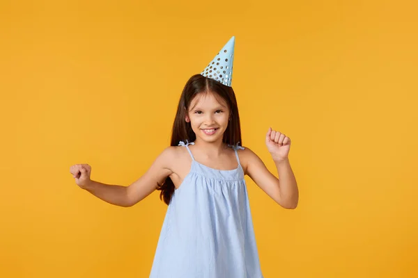 happy birthday child girl in blue dress and party hat dancing on yellow background