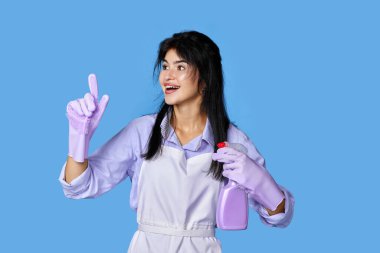 Menekşe rengi lastik eldivenli ve önlüklü güzel bir kadın. Deterjan spreyli, işaret parmağıyla mavi arka planda izole edilmiş yeni bir fikir tutuyor. temizlik