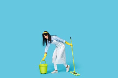 Lastik eldivenli bir kadın ve temiz önlüklü bir kadın mavi arka planda paspasla yerleri yıkıyor. Tam uzunluk